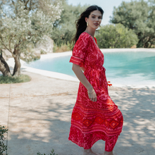 Load image into Gallery viewer, Woman in a red dress standing by a pool with trees in the background