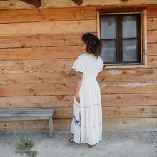 Load image into Gallery viewer, Woman in a white dress standing in front of a wooden cabin