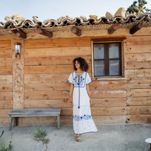 Load image into Gallery viewer, Woman in a white dress with blue embroidery standing in front of a wooden cabin.