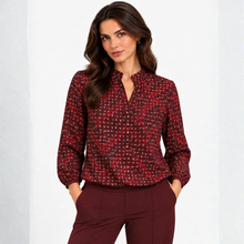 Load image into Gallery viewer, A woman modeling a long-sleeve blouse with a red and black micro-tweed pattern and a split V-neckline, paired with burgundy trousers against a light gray studio background.