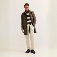 Load image into Gallery viewer, A male model stands against a plain off-white studio background wearing a brown mid-length overcoat over a brown and cream striped knit sweater, cream trousers, a black belt, and dark brown leather shoes.