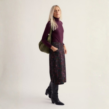 Load image into Gallery viewer, A woman stands in a studio wearing a dark purple turtleneck, a dark patterned midi skirt with pockets, black knee-high boots, and carrying an olive green shoulder bag.