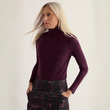 Load image into Gallery viewer, A blonde woman wearing a fitted long-sleeve turtleneck top in deep plum purple, paired with a dark patterned skirt, standing against a plain off-white background.