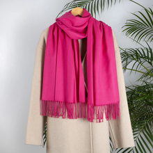 Load image into Gallery viewer, A bright pink scarf with fringe trim draped over a beige coat on a wooden hanger, set against a light gray wall with green palm leaves in the background.