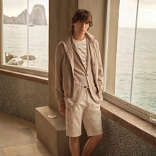 Load image into Gallery viewer, Person wearing a beige jacket and shorts standing by large windows with a scenic view.