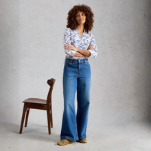 Load image into Gallery viewer, Woman wearing a floral blouse and blue jeans standing against a plain background with a wooden chair.
