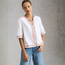 Load image into Gallery viewer, Woman wearing a white blouse with red patterns and blue jeans against a gray background