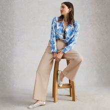 Load image into Gallery viewer, Woman sitting on a stool wearing a blue floral blouse and beige pants against a plain background