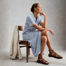 Load image into Gallery viewer, Woman sitting on a chair wearing a blue patterned dress and brown sandals, with a neutral background.