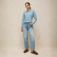 Load image into Gallery viewer, A woman modeling a blue and white striped long-sleeve knit top with light-wash blue jeans and brown suede loafers, standing against a neutral textured beige backdrop.