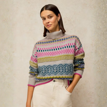 Load image into Gallery viewer, A woman wearing a multicolor Fair Isle patterned turtleneck sweater with pink, blue, yellow, and gray tones, paired with cream-colored pants, standing against a textured beige wall.