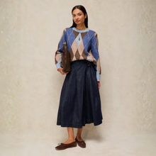 Load image into Gallery viewer, A woman stands against a textured beige studio wall wearing a blue, beige, and brown argyle sweater, a dark navy midi skirt, and brown loafers, holding a dark brown clutch.