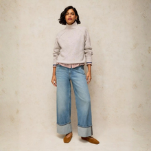 Load image into Gallery viewer, A woman stands against a textured light grey studio wall wearing a light beige knit mock-neck sweater, wide-leg light blue jeans with rolled cuffs, and tan flats.