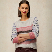 Load image into Gallery viewer, A woman wearing a heather gray knit sweater with horizontal stripes in dusty pink, dark brown, and light blue, paired with dark brown pants, standing against a textured beige wall.