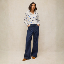 Load image into Gallery viewer, A woman stands against a textured beige studio backdrop wearing a white long-sleeve button-up shirt with black and blue polka dots, dark wide-leg denim jeans, and tan pointed shoes.