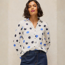 Load image into Gallery viewer, A woman wearing a white long-sleeve button-up blouse with blue and black polka dots, paired with dark blue jeans, standing against a neutral beige wall.