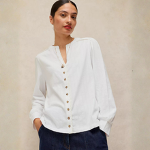 Load image into Gallery viewer, A woman wearing a white long-sleeve button-front blouse with scalloped trim and dark blue jeans, standing against a plain beige wall.