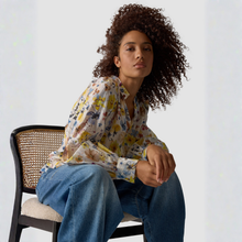 Load image into Gallery viewer, Woman sitting on a chair wearing a floral blouse and blue jeans against a plain background