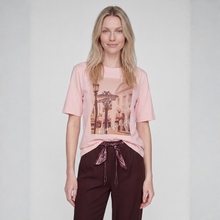 Load image into Gallery viewer, A woman modeling a light pink short-sleeve graphic t-shirt with a vintage street scene print, paired with dark brown drawstring pants, against a light gray studio background.