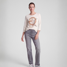 Load image into Gallery viewer, A woman modeling a cream-colored long-sleeve top with a large metallic bronze circular logo on the front, paired with gray jeans and white sneakers, against a light gray studio background.
