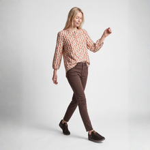 Load image into Gallery viewer, A woman modeling a long-sleeve blouse with a peach and brown geometric pattern, paired with brown pants and dark brown sneakers, against a light gray studio background.