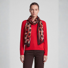 Load image into Gallery viewer, A woman modeling a long scarf with an abstract chain-link pattern in black, rust orange, and cream, worn over a bright red sweater and dark brown trousers against a light gray studio background.