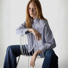 Load image into Gallery viewer, A woman with long red hair sits on a white wire chair, wearing a blue and white vertically striped button-down shirt and dark blue jeans against a plain white background.