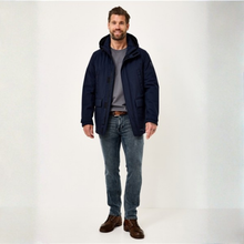 Load image into Gallery viewer, A man wearing a navy blue hooded jacket over a gray t-shirt, blue jeans, and brown shoes, standing against a plain white studio background.
