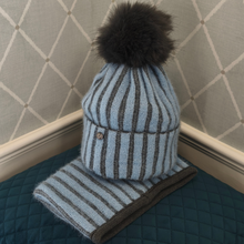 Load image into Gallery viewer, A cosy light blue knit hat topped with a statement black faux-fur pom-pom, accompanied by a matching striped scarf for a coordinated winter look. Presented on a blue quilted cushion against a crisp white panelled wall.