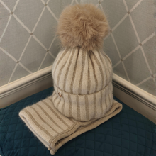 Load image into Gallery viewer, A stylish beige knitted beanie featuring a large, fluffy brown faux-fur pom-pom, paired with a coordinating striped knit scarf. Beautifully displayed on a blue quilted cushion against a white panelled backdrop.