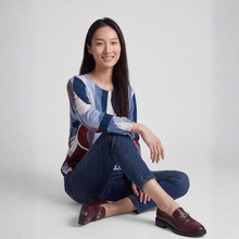 Load image into Gallery viewer, A woman sits on the floor wearing a long-sleeve abstract print blouse in shades of blue, burgundy, and cream, paired with dark blue jeans and burgundy leather loafers with gold hardware, against a light gray studio background.