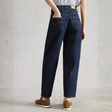 Load image into Gallery viewer, A person shown from the back wearing dark blue straight-leg jeans, a striped top, and brown suede shoes, standing against a textured light gray wall.