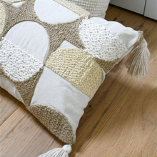 Load image into Gallery viewer, A square decorative throw pillow with a patchwork design of textured knit and woven panels in beige, cream, and white, featuring tassels on the sides, resting on a light brown wooden floor.