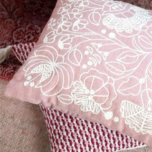 Load image into Gallery viewer, A square pink throw pillow with white floral embroidery, resting on top of a textured red and white woven cushion.