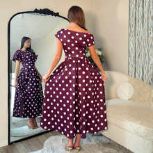Load image into Gallery viewer, A woman stands in a living room wearing a burgundy off-the-shoulder midi dress with white polka dots, holding the skirt out to show its shape. A large black-framed mirror on the left reflects her profile, and a cream-colored sofa is visible on the right.