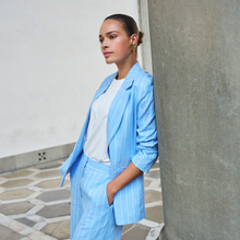Load image into Gallery viewer, Woman in a light blue suit standing against a gray wall.