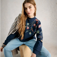 Load image into Gallery viewer, Woman wearing a navy sweater with floral embroidery sitting on a wooden stool.