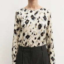 Load image into Gallery viewer, A woman wearing a long-sleeve blouse with an abstract leopard-like print in black, brown, and muted blue-gray on an off-white base, paired with dark charcoal trousers, against a plain light background.
