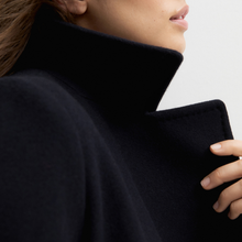 Load image into Gallery viewer, Close-up of a person wearing a dark navy or near-black wool coat with a very high structured collar, showing part of the face, hand, and shoulder against a light neutral background.