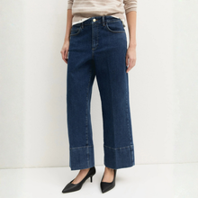 Load image into Gallery viewer, A model is wearing dark blue wide-leg cropped jeans with rolled cuffs, paired with a beige striped top and black pointed-toe heels, against a plain white studio background.