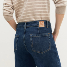 Load image into Gallery viewer, Rear view of a person wearing dark blue denim jeans with a tan square patch featuring the letters 'M' and 'A' on the back waistband, paired with a beige and white striped long-sleeve top against a light off-white background.