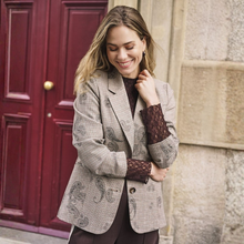 Load image into Gallery viewer, A woman wearing a beige and brown patterned blazer over a dark brown lace-sleeve top, standing outdoors in front of a red wooden door and a stone wall.