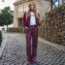 Load image into Gallery viewer, A woman stands on a cobblestone street wearing a burgundy bomber jacket, a white graphic T-shirt, and shiny burgundy wide-leg pants, with matching burgundy shoes. A stone building and stone wall are in the background.