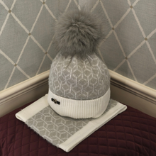 Load image into Gallery viewer, A stylish knitted beanie featuring a white and grey geometric pattern, a cosy white ribbed cuff and a large grey faux-fur pom-pom. Displayed on a burgundy quilted cushion against a grey diamond-patterned background.