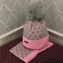Load image into Gallery viewer, A cosy knitted beanie hat featuring a stylish pink and grey geometric design, complemented by a soft pink ribbed cuff and a large grey faux-fur pom-pom. Beautifully displayed on a burgundy quilted cushion against a grey diamond-patterned backdrop.