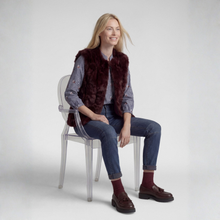 Load image into Gallery viewer, A woman sits on a clear acrylic chair wearing a dark burgundy faux fur vest over a blue and white striped button-down shirt, blue jeans, burgundy socks, and brown leather loafers, against a light gray studio background.