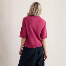 Load image into Gallery viewer, A woman viewed from behind wearing a short-sleeve knit sweater in a rich magenta-pink color, paired with dark navy trousers, against a plain off-white background.