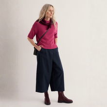 Load image into Gallery viewer, A woman modeling a short-sleeve magenta knit sweater with a dark cowl neck, paired with dark navy wide-leg cropped pants and burgundy lace-up ankle boots, against a plain off-white studio background.