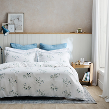 Load image into Gallery viewer, A neatly made bed in a bright bedroom featuring a white duvet cover and matching pillowcases with a delicate blue and green floral print. The bed is styled with two solid blue pillows, set against a white paneled headboard with a light wood shelf, a small wooden side table, and a textured beige wall.
