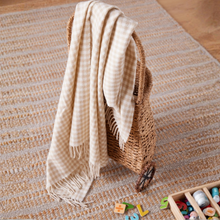 Load image into Gallery viewer, A beige and cream gingham check throw blanket with fringe is draped over a woven wicker basket on a striped woven rug.